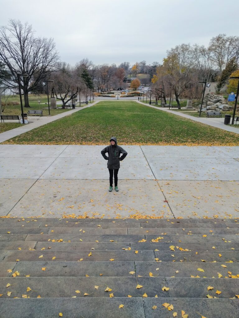 Maggie stands with hands on her hips, waiting for the next round of stairball. Behind her a lawn stretches towards a traffic circle and trees in the distance.