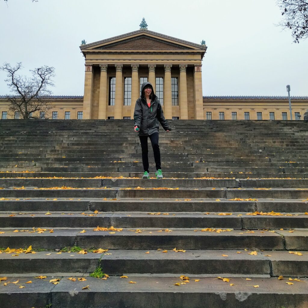 A person with a maniacal grin is preparing to toss a racquetball down the stairs behind the Philadelphia Museum of Art. It looks cold and wintry, with leaves on the ground. She is wearing a jacket and leggings.